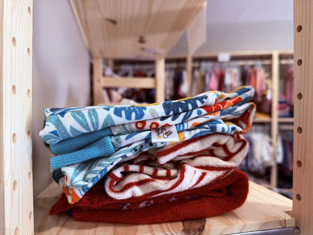 Pile de vêtements enfants d’occasion pliés dans une boutique de seconde main avec portants en arrière-plan